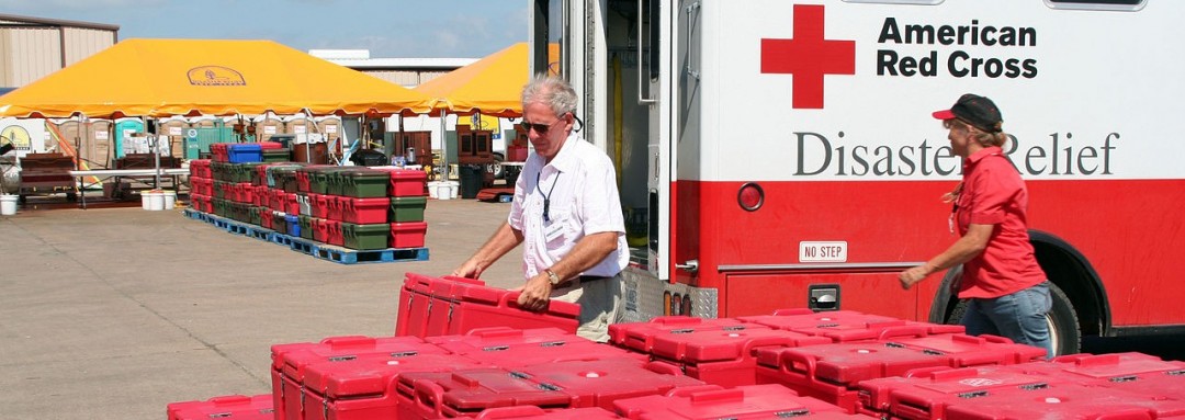 Red Cross staff in front of a Red Cross ambulance, red plastic food containers in foreground